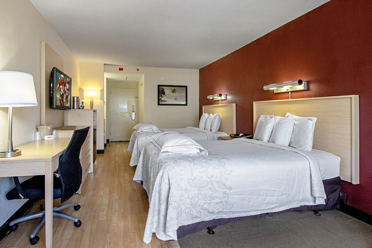 A hotel room with three beds, a desk with a chair, a lamp, and a painting on the wall.