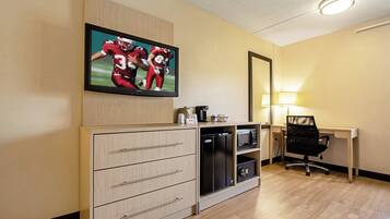 A hotel room with a flat-screen TV mounted on the wall, a desk with a chair, a microwave, and a refrigerator.