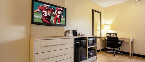A hotel room with a flat-screen TV mounted on the wall, a desk with a chair, a microwave, and a refrigerator.
