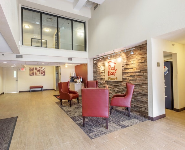 Un hall d'entrée avec une enseigne « Red Roof Plus », un espace détente avec des fauteuils, un comptoir d'accueil et un escalier menant à un étage supérieur.