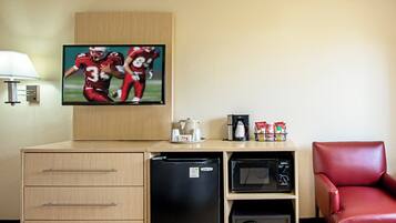 A hotel room with a television, a coffee maker, a microwave, a refrigerator, a red chair, and a wooden cabinet with drawers.