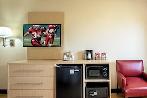 Een hotelkamer met een televisie, koffiezetapparaat, magnetron, koelkast, een rode stoel en een houten dressoir met lades.