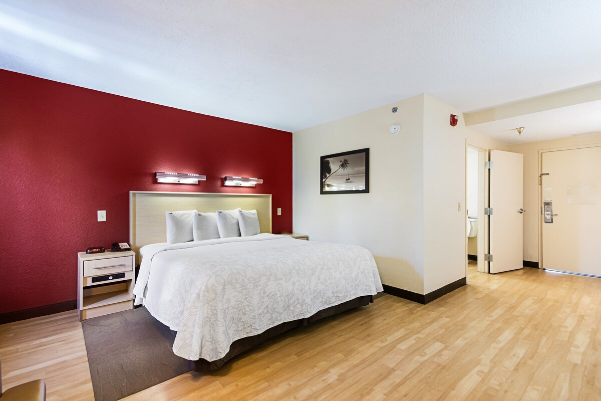 A hotel room with a bed, bedside tables, a nightstand, a red wall, a white wall, a framed picture, and a door.