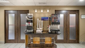 Zona de recepción de hotel con estación de café, barra de zumos y mostrador con un cartel que dice 'Hydrate Juice Bar'.