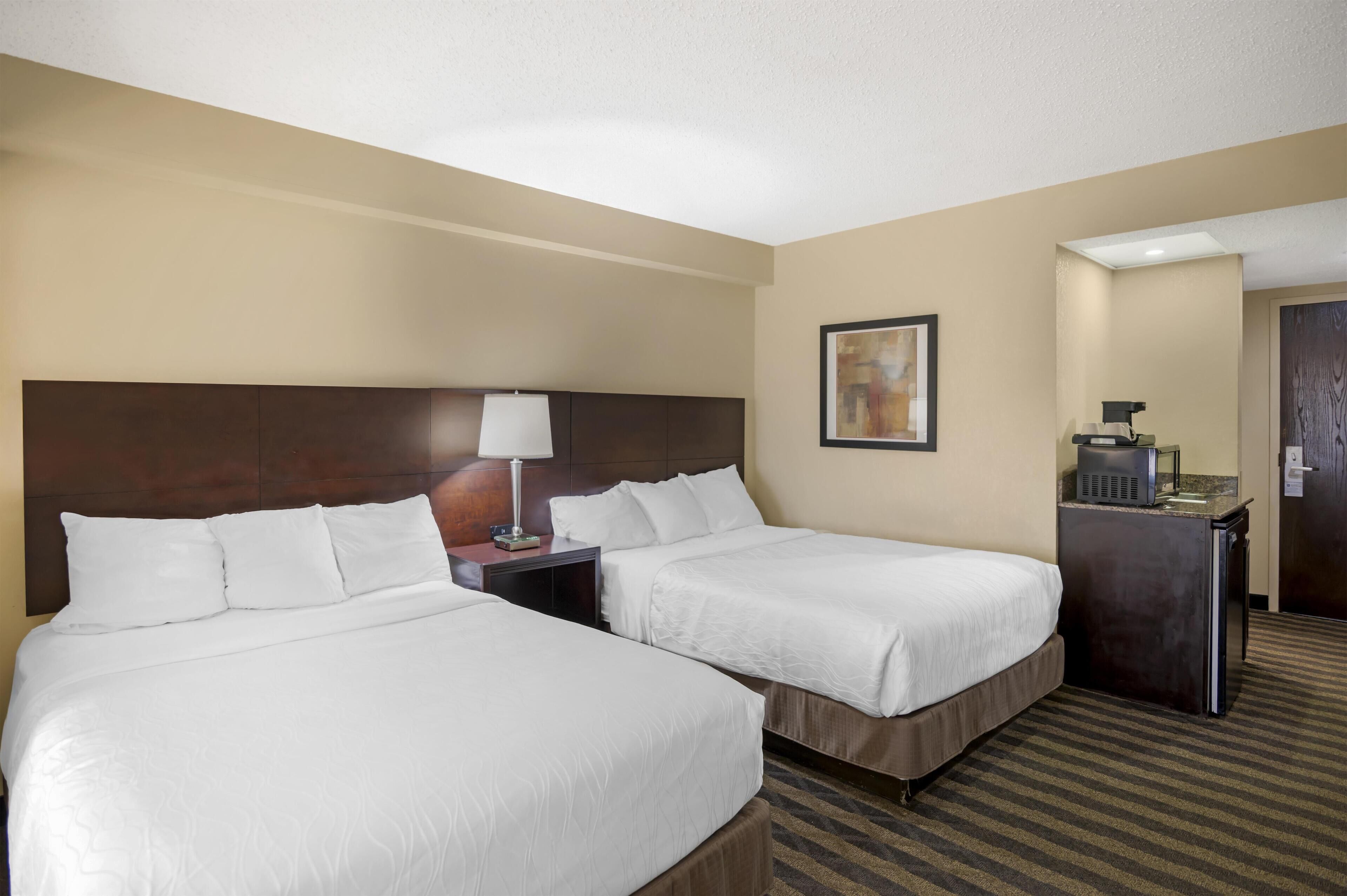 A hotel room with two beds, a desk, a lamp, a painting, and a minibar.