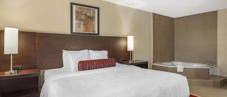 Habitación de hotel con una cama grande, dos mesitas de noche con lámparas, un cuadro en la pared y un baño separado con bañera.