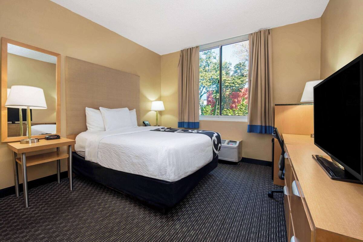 A hotel room with a bed, a desk with a lamp, a television, and a window with curtains.