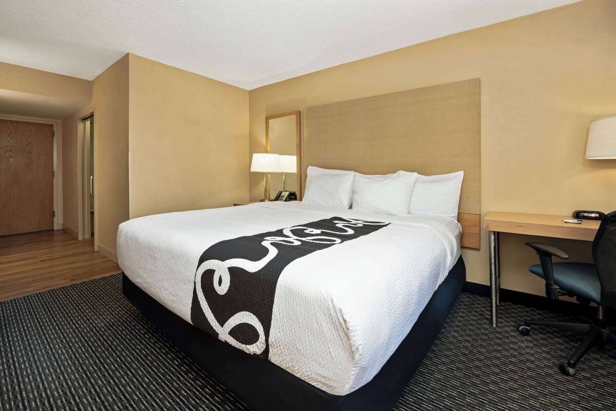 A hotel room with a large bed, a desk, a chair, a lamp, and a door.