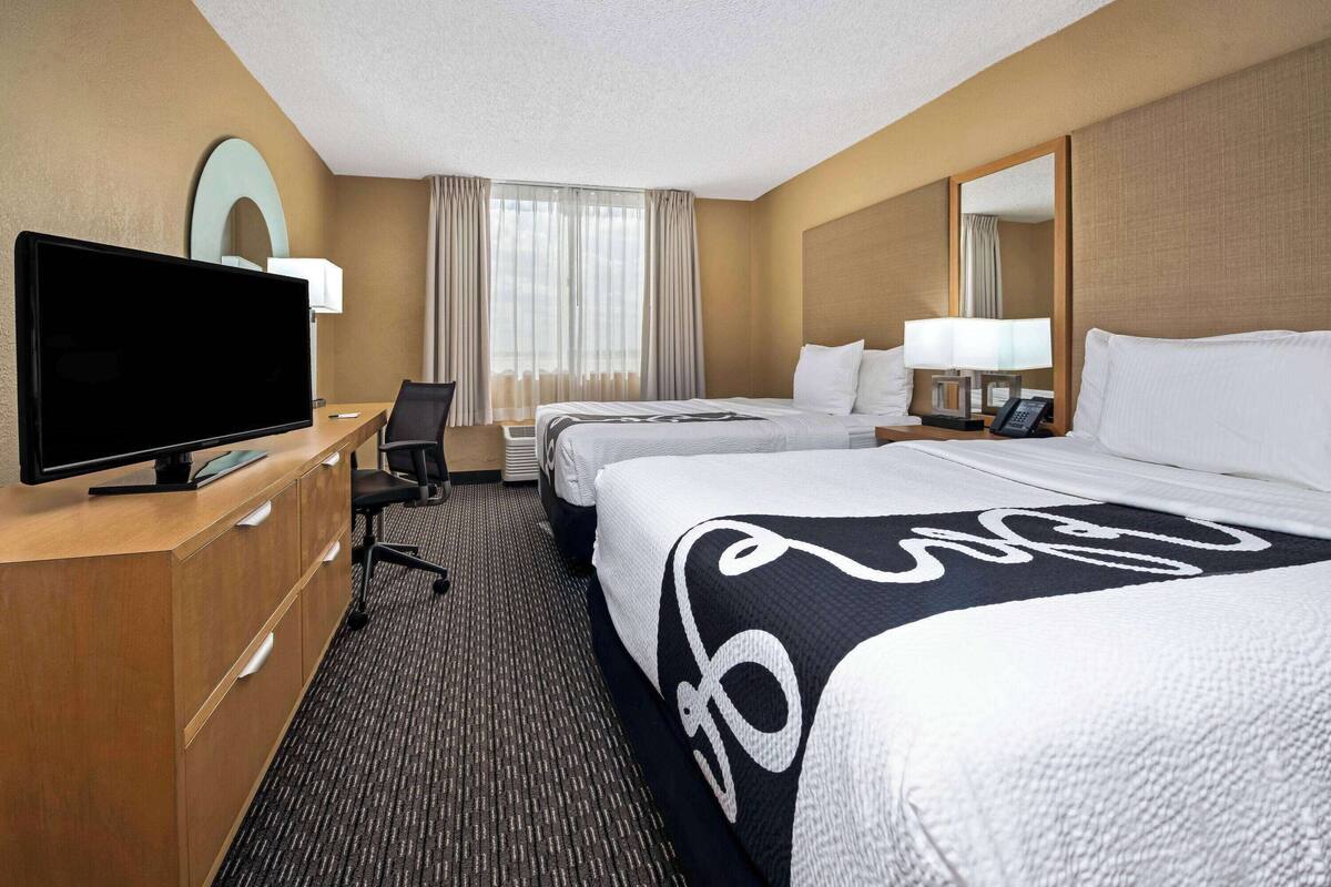 A hotel room with a flat-screen TV, two beds, a desk with a chair, and a large mirror.