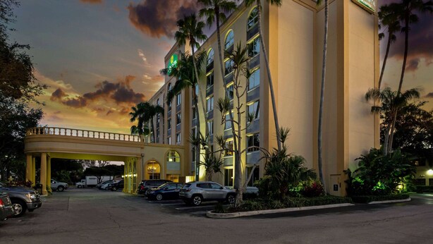A hotel with a central entrance, multiple floors, and palm trees. - La Quinta Inn & Suites by Wyndham West Palm Beach Airport (West Palm Beach)