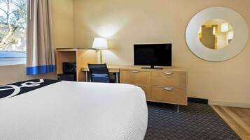 Habitación de hotel con una cama grande, un escritorio con computadora, televisor, espejo y ventana con cortinas.
