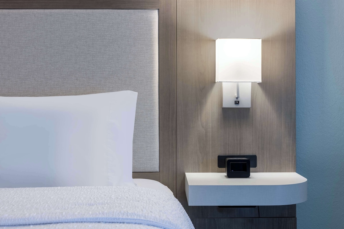 A hotel room with a bed, a nightstand, a wall-mounted lamp, and a power outlet.