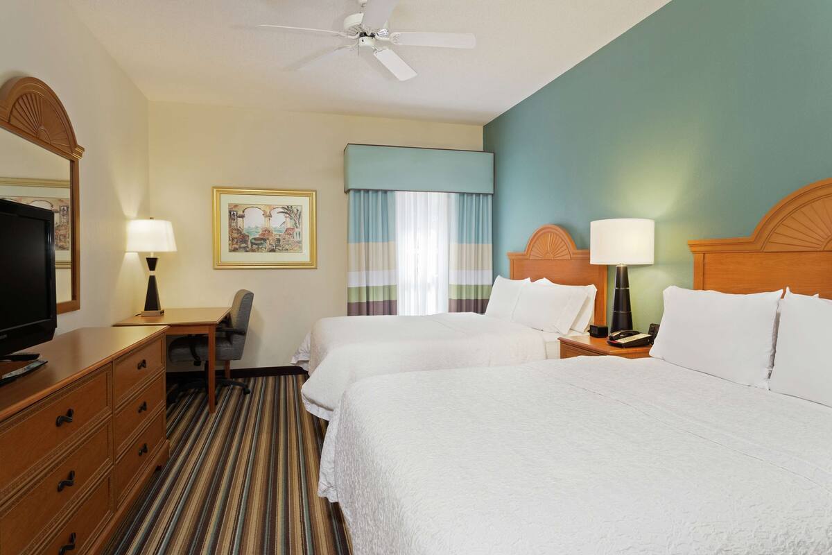 A hotel room with two beds, a desk, a chair, a television, and a ceiling fan.
