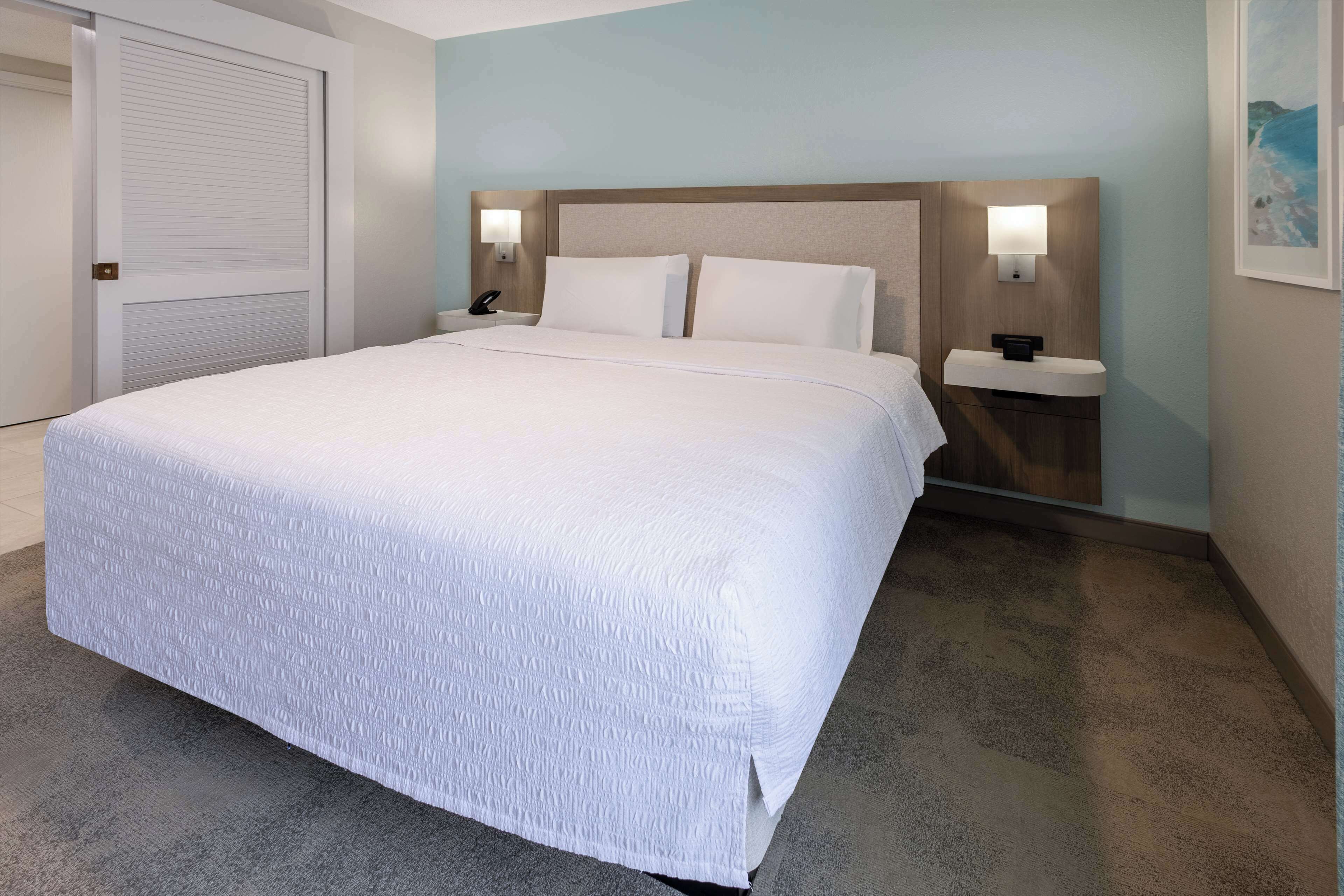 A hotel room with a large bed, two bedside tables with lamps, a door, and a framed picture on the wall.