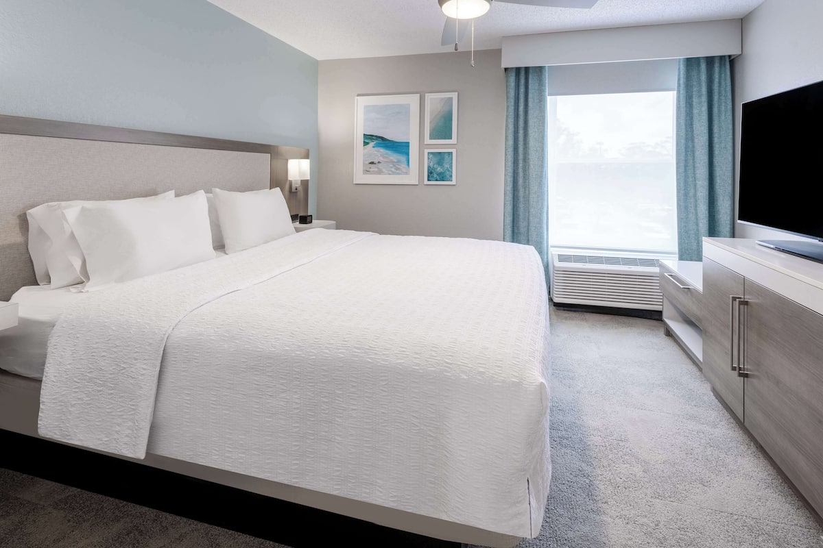 A neatly made bed with white linens, a flat-screen TV, a bedside table with a lamp, and a large window with curtains.