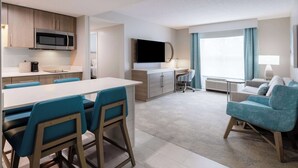 Moderni hotellihuone, jossa on keittotila, ruokapöytä tuoleineen, sohva ja televisio.