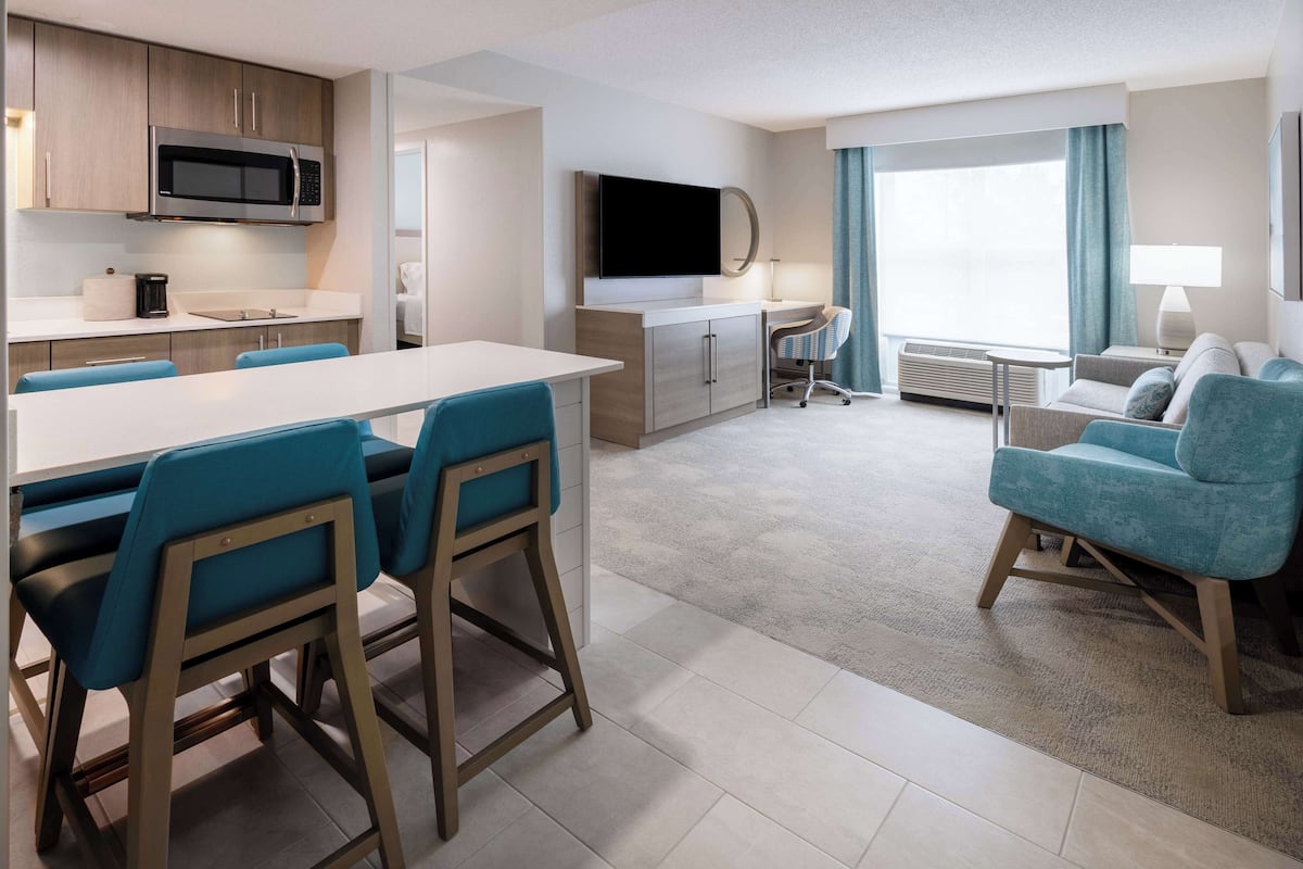A modern hotel room with a kitchen area, a dining table with chairs, a sofa, and a TV.