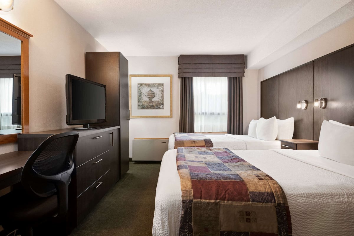 A hotel room with a desk, television, two beds, a large window with curtains, and a framed picture on the wall.