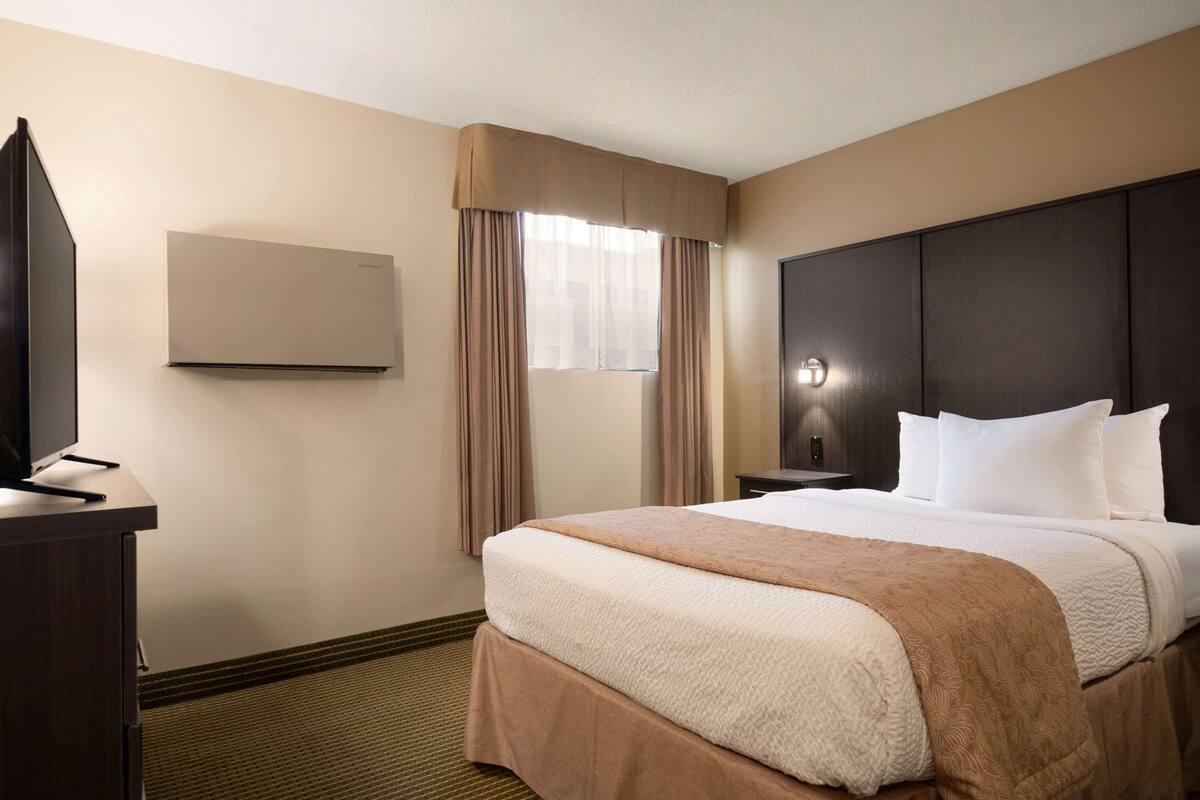 A hotel room with a large bed, a flat-screen TV, a wall-mounted air conditioner, and a window with curtains.