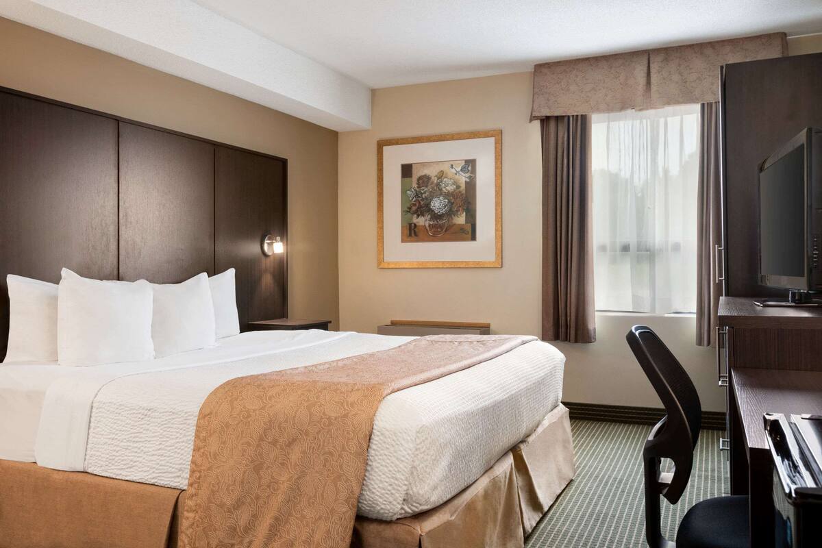 A hotel room with a large bed, a desk with a computer, a chair, a television, and a framed artwork on the wall.