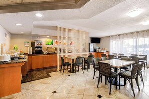 A hotel dining area with a kitchen in the background, a counter with a beverage dispenser, and a seating area with tables and chairs.