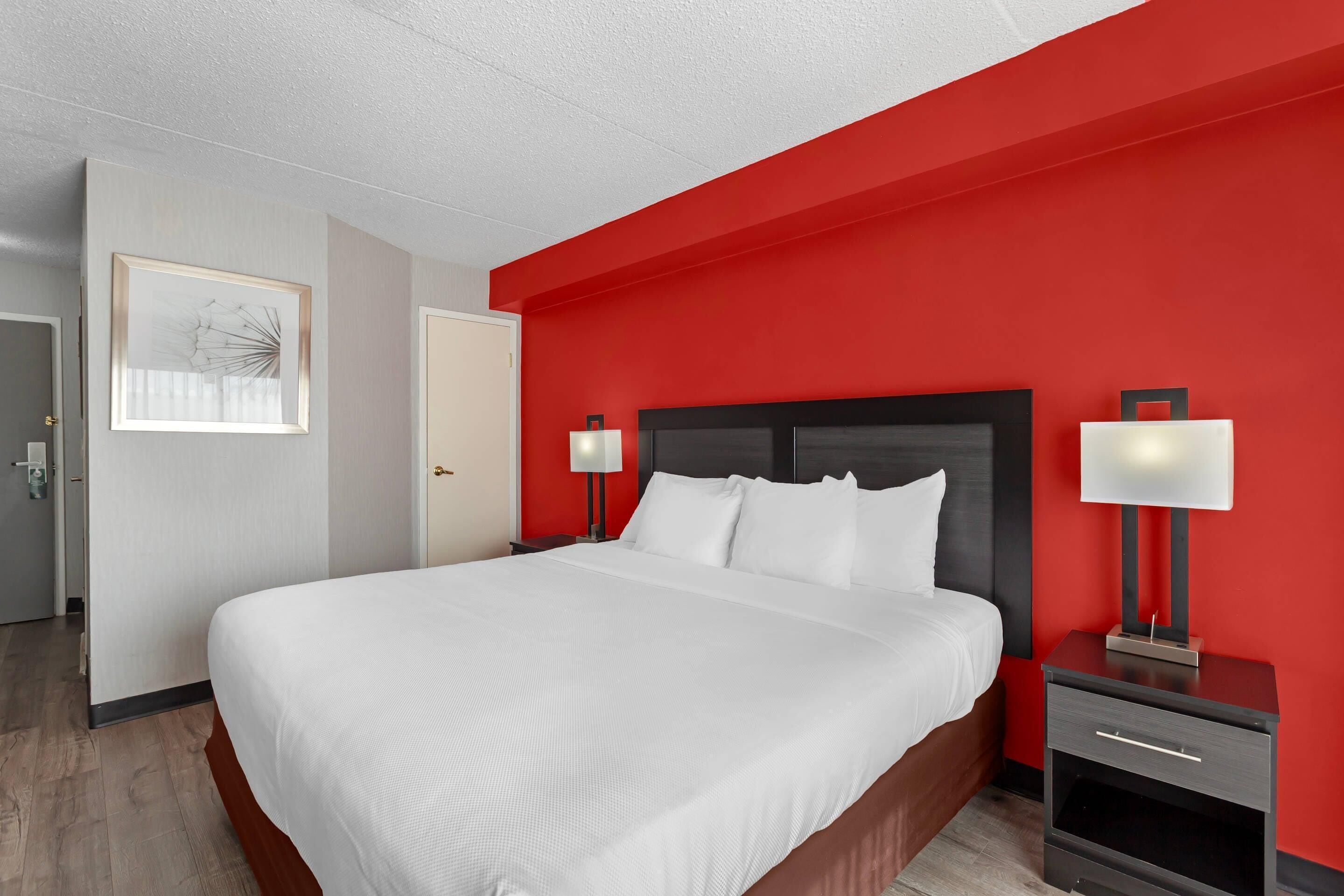A hotel room with a large bed, two bedside tables with lamps, a framed artwork on the wall, and a door leading to another room.