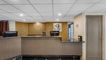 A hotel reception area with a wooden reception desk, a sign that reads "Quality," and a door to the right.