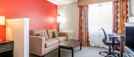A hotel room with a sofa, a coffee table, a desk with a chair, a lamp, and a television.