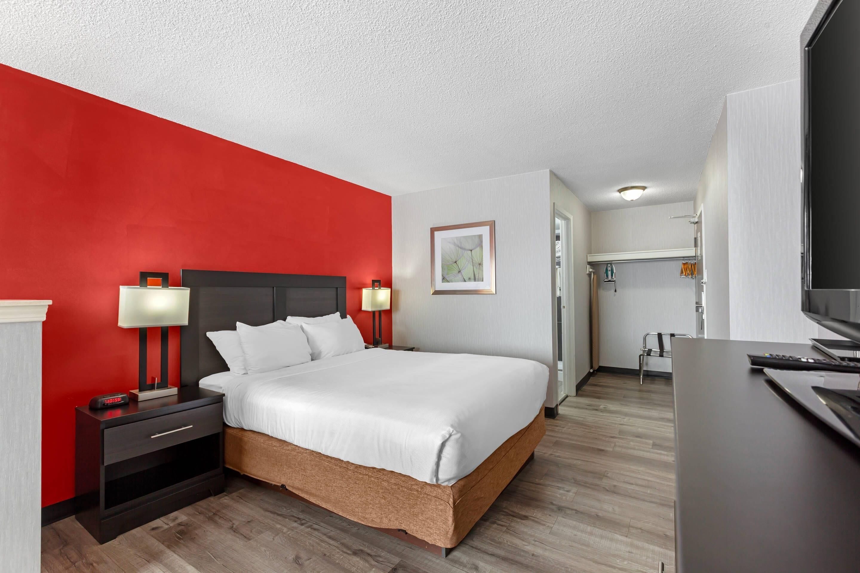 A hotel room with a red accent wall, a bed with white linens, a nightstand with a lamp, a desk with a flat-screen TV, and a door leading to a bathroom.