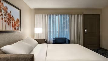 Quarto de hotel com uma cama grande, uma cadeira, uma escrivaninha, um abajur e uma porta.