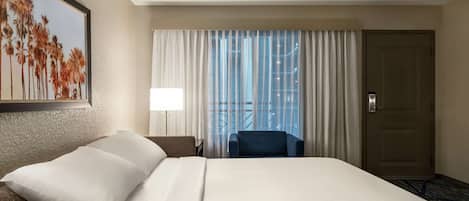 A hotel room with a large bed, a chair, a desk, a lamp, and a door.