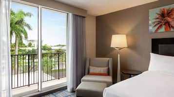 A hotel room with a large window, a bed, a chair, a lamp, and a painting of a palm tree.