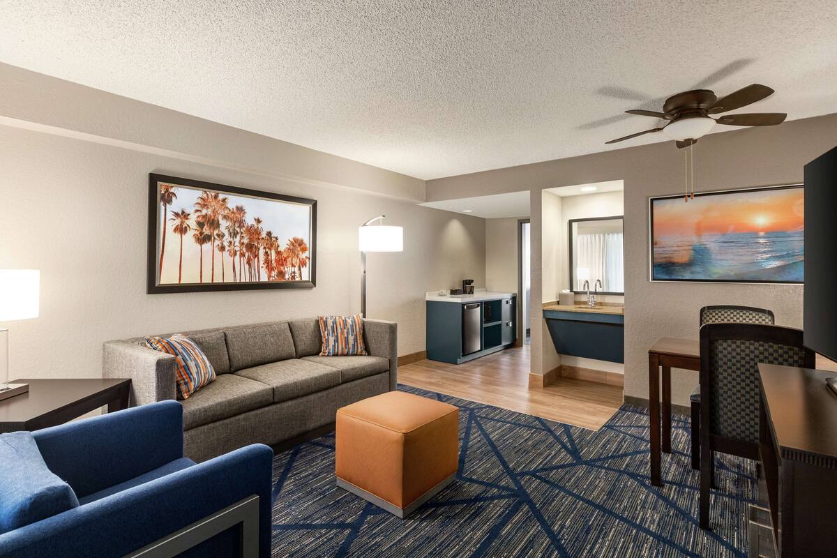 Een moderne hotelkamer met een bank, poef, eettafel en stoelen. Achterin is een kitchenette en er hangt een plafondventilator.