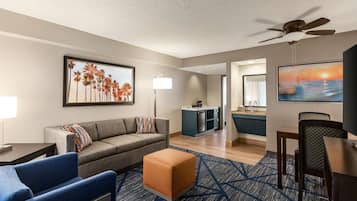 Habitación de hotel moderna con sofá, reposapiés, mesa de comedor y sillas. Se observa una kitchenette al fondo y un ventilador de techo.