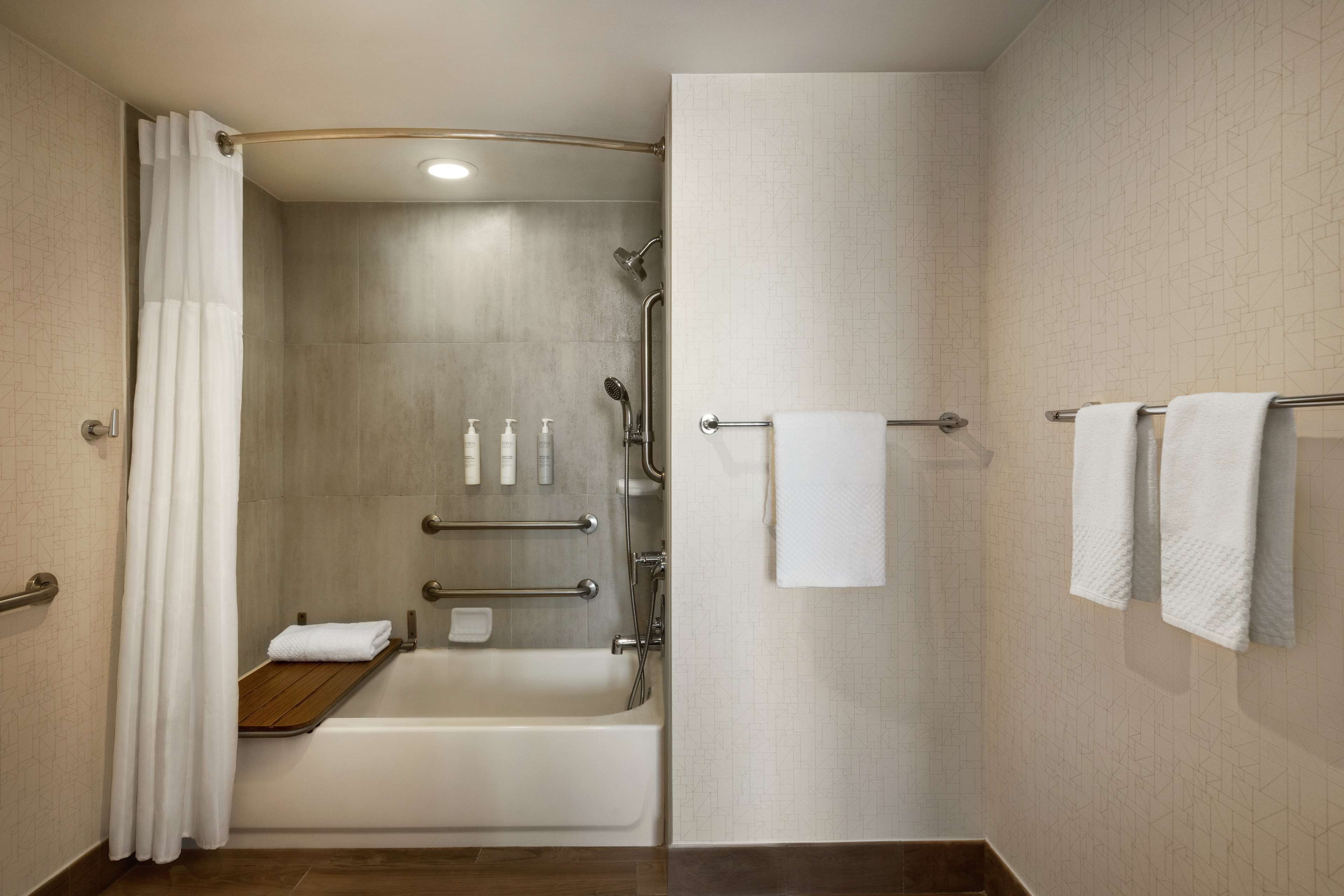 A modern bathroom with a bathtub, shower, and handrails. There are towels and toiletries visible.