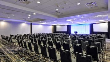 Un salón de conferencias con filas de sillas, una pantalla grande y un proyector.