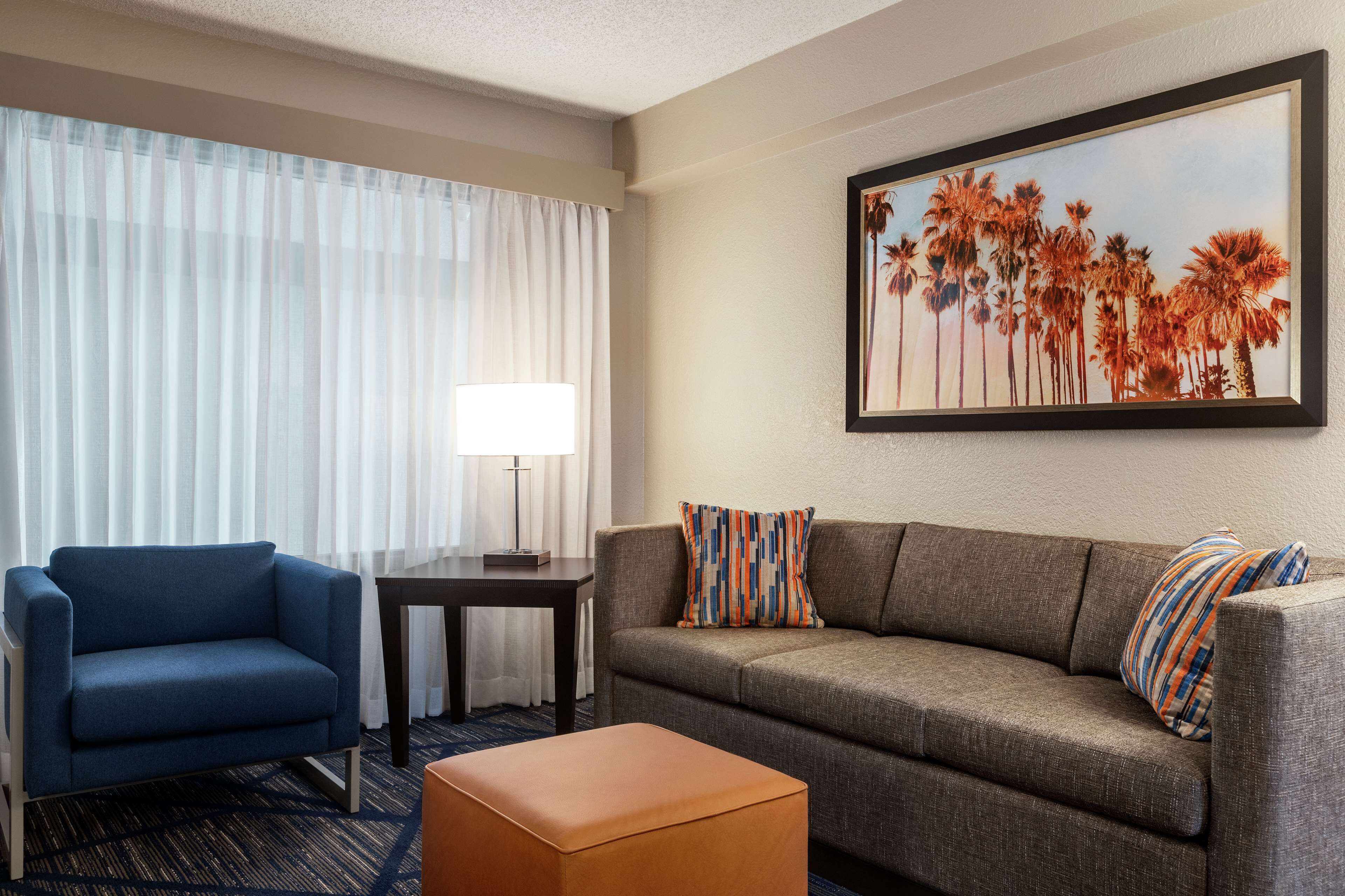 A hotel room with a blue armchair, a gray sofa with striped pillows, a small table with a lamp, and a framed picture of palm trees on the wall.