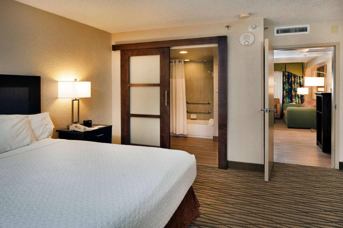 Een hotelkamer met een bed, een bedlampje, een badkamer met douche, en een groene bank in de aangrenzende kamer.