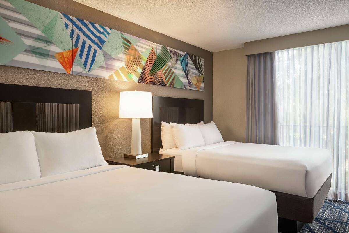 Een hotelkamer met twee bedden, een groot abstract schilderij aan de muur, een lamp en een raam met gordijnen.
