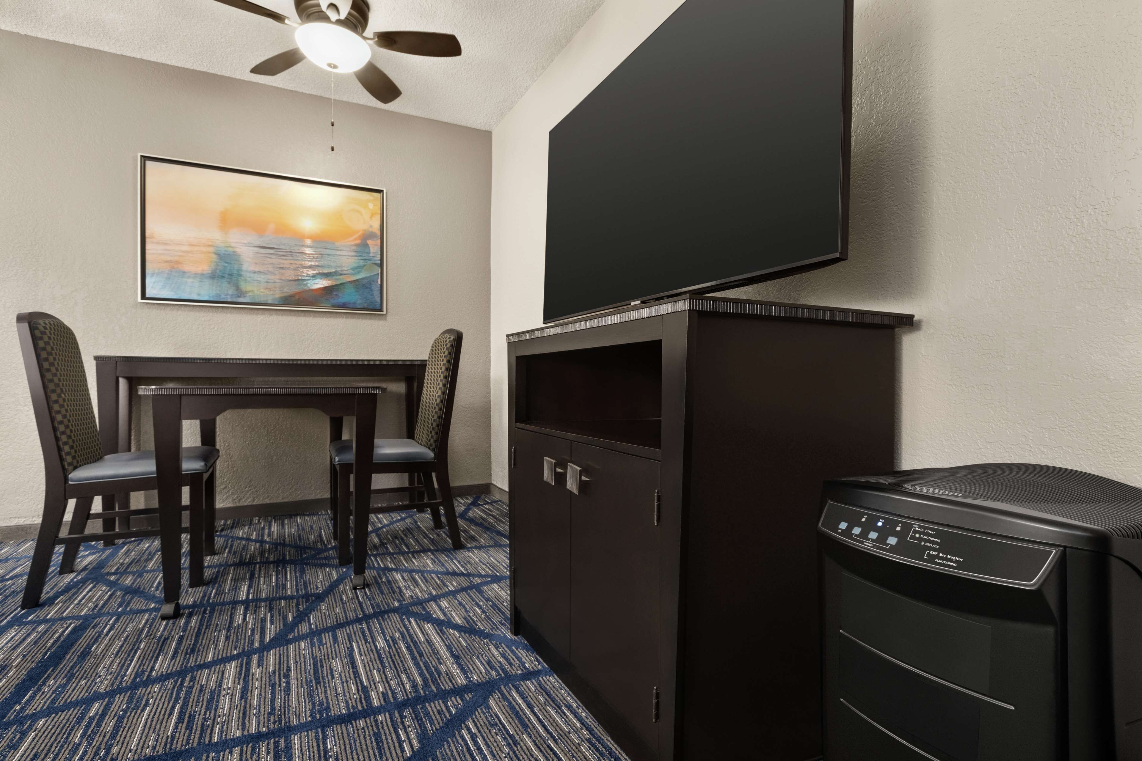Habitación de hotel con televisor de pantalla plana montado en la pared, un mueble bajo con panel de control, una mesa de comedor con sillas y piso alfombrado.
