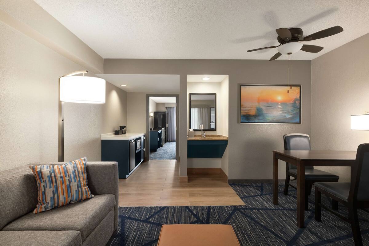 Een hotelkamer met een slaapbank, een eettafel, een plafondventilator, een kitchenette en een schilderij aan de muur.