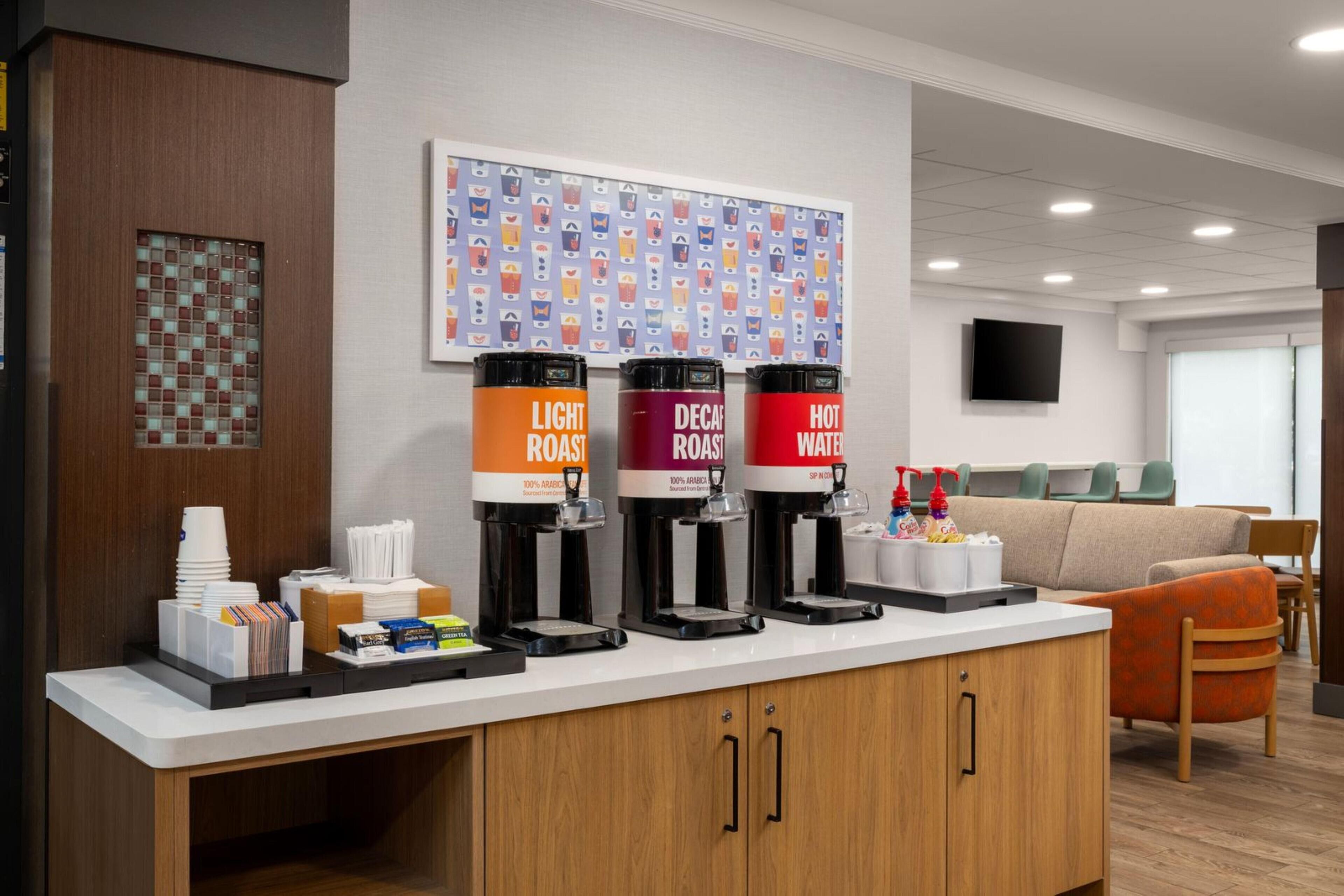 A coffee station with three coffee dispensers labeled "Light Roast," "Decaf Roast," and "Hot Water." There are stacks of white cups and a small table with condiments and a bottle of hand sanitizer.