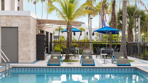 Une piscine entourée de palmiers et d’un bâtiment moderne, avec des chaises longues et des parasols à proximité.