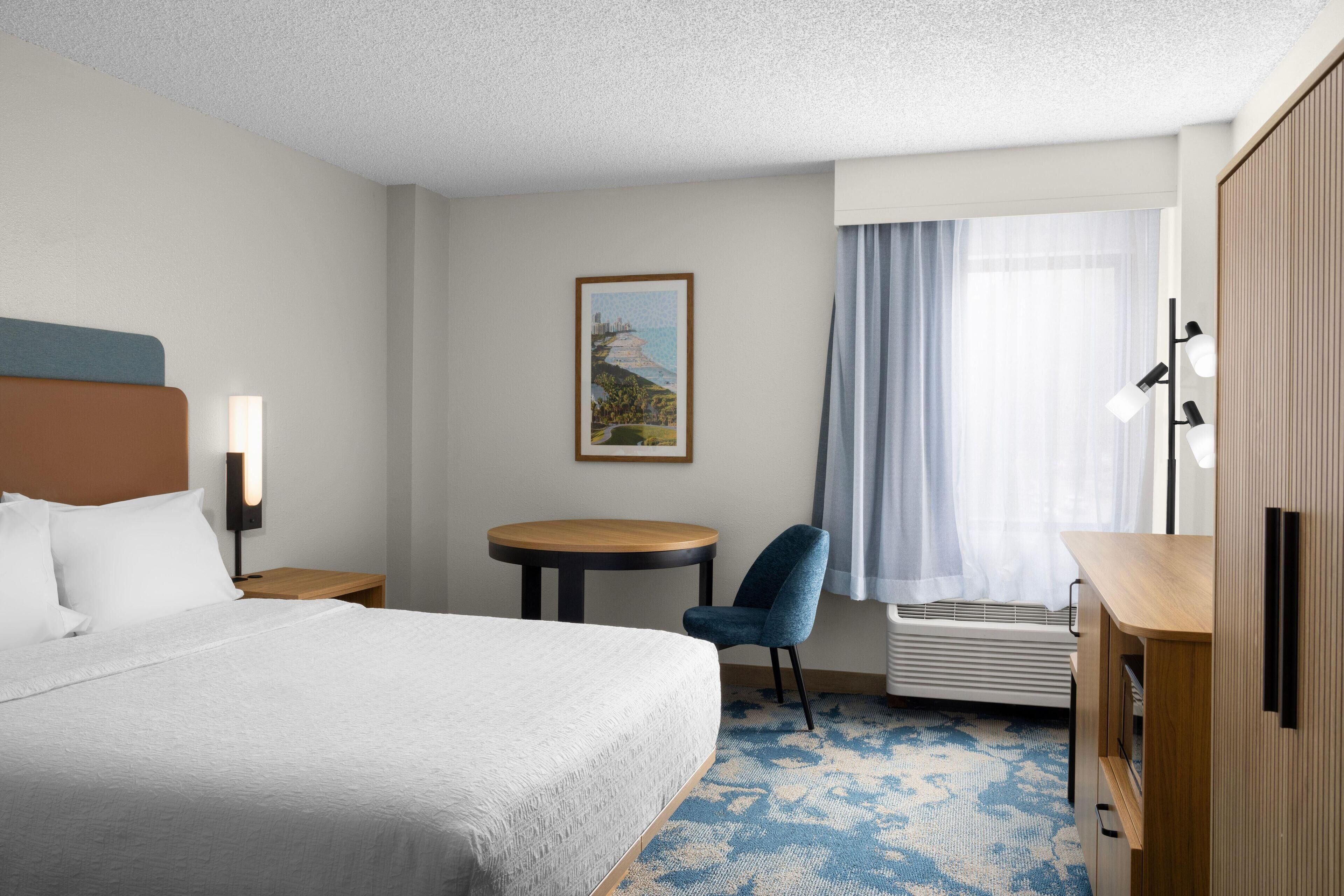 Habitación de hotel con cama, escritorio, silla, una mesita, un cuadro en la pared y ventana con cortinas.