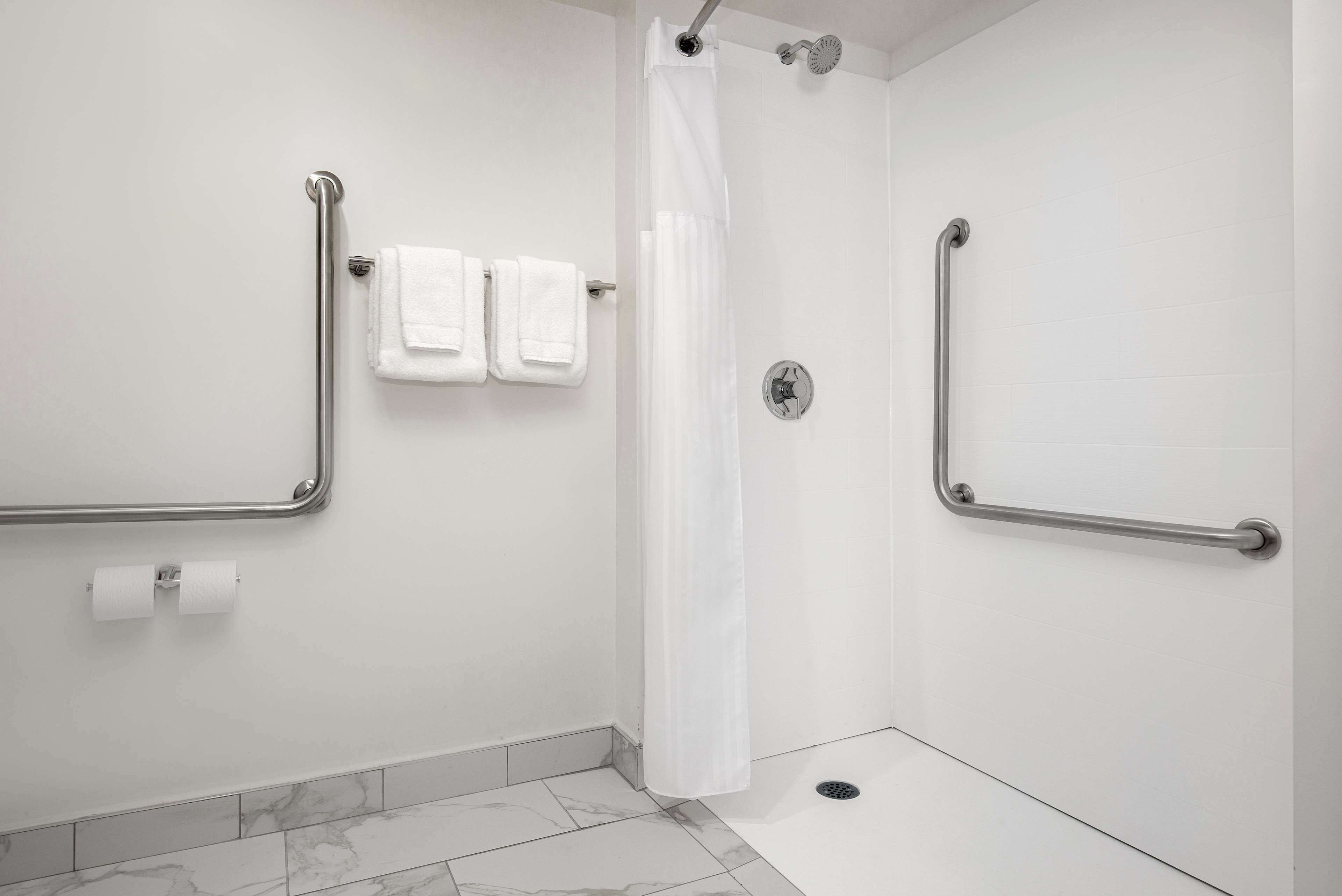Un bagno con pareti bianche, una doccia con tenda bianca e due maniglie di sostegno a parete.