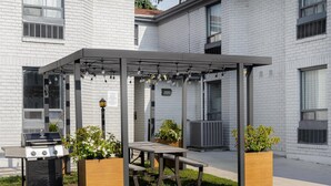 An outdoor patio with a grill, wooden table, and potted plants. - Spark by Hilton Brockville (Brockville)