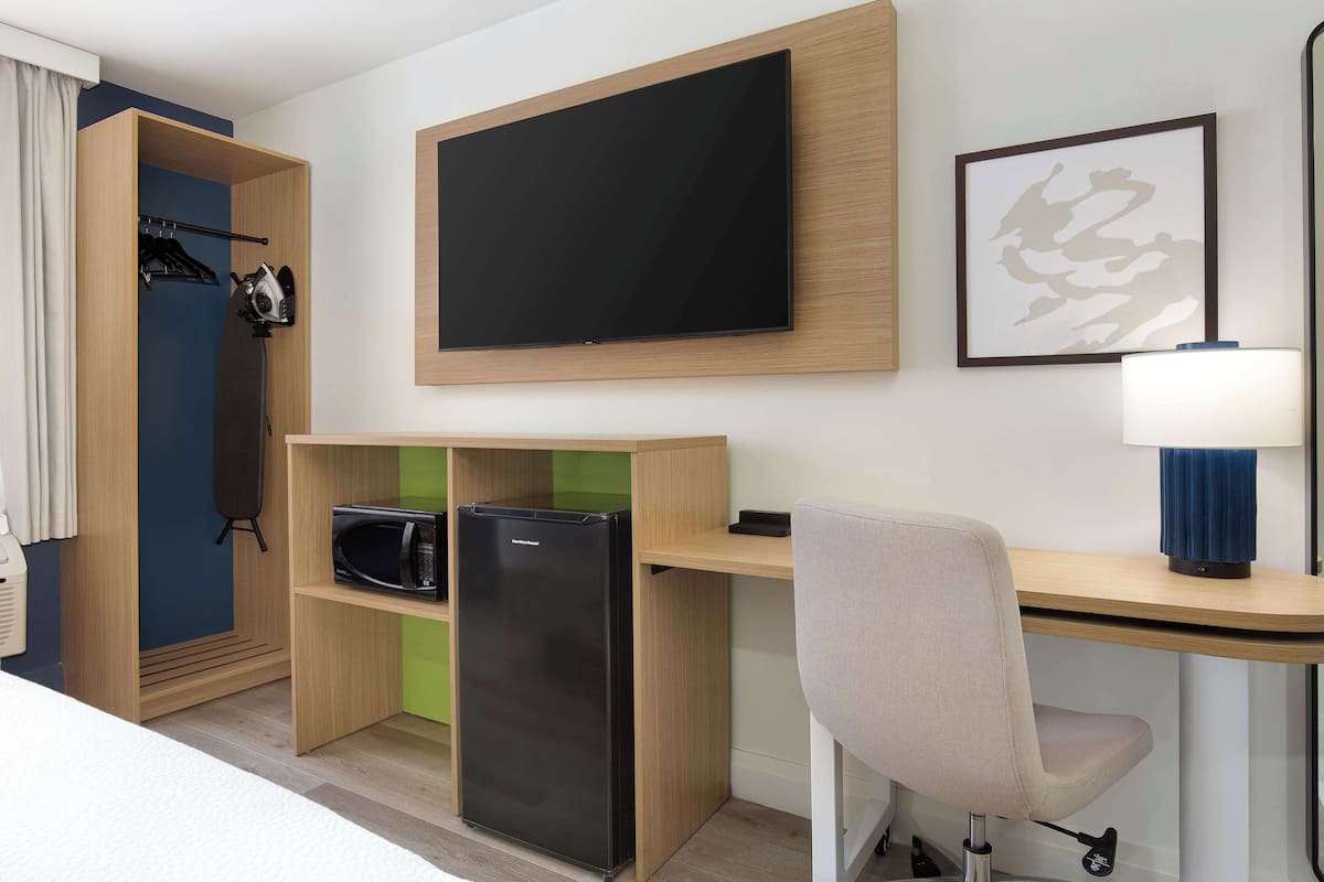 A compact hotel room with a bed, a desk with a computer, a flat-screen TV, a microwave, and a refrigerator.