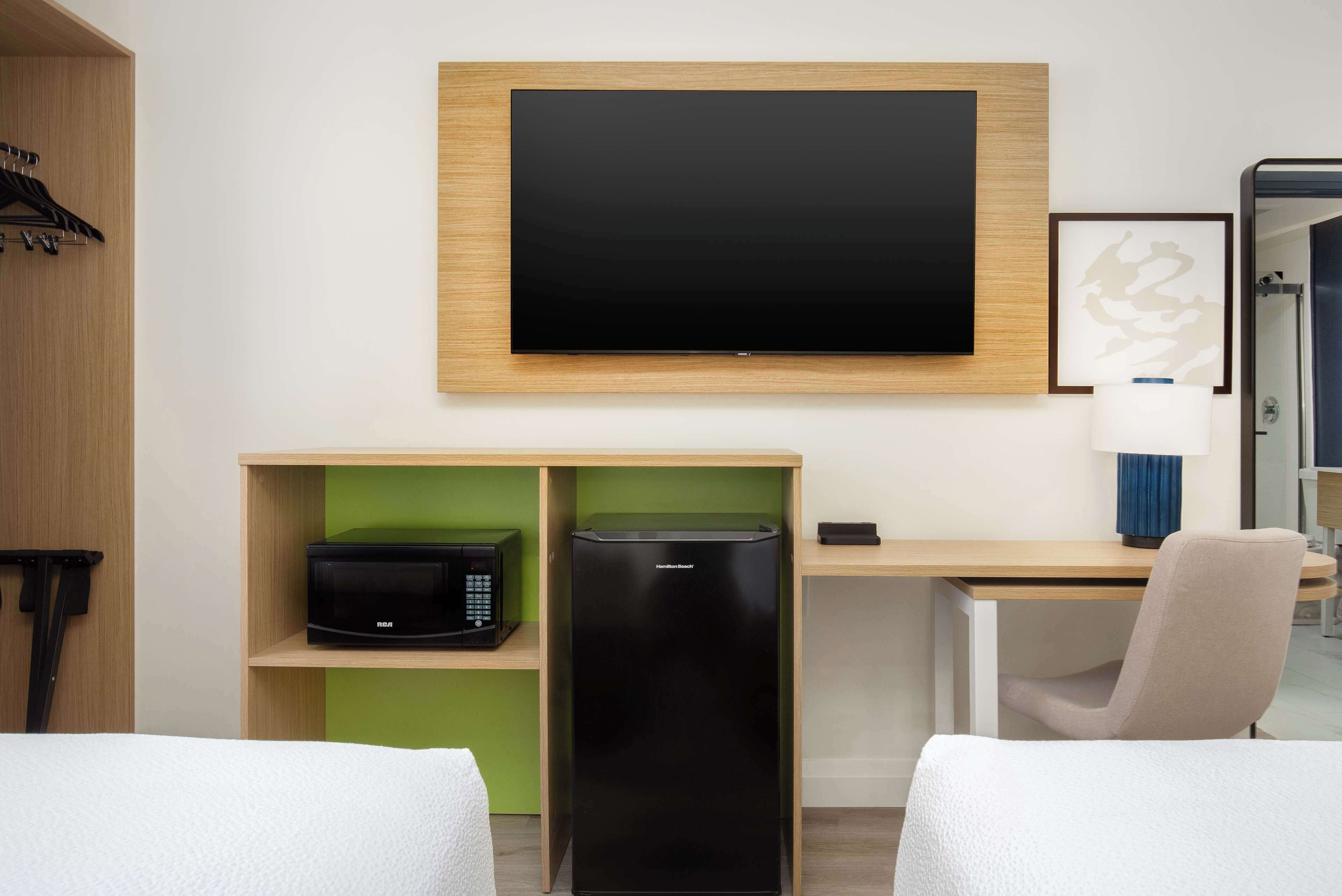 A hotel room with a flat-screen TV mounted on the wall, a microwave, a mini-fridge, a desk with a chair, and a bed with a white comforter.