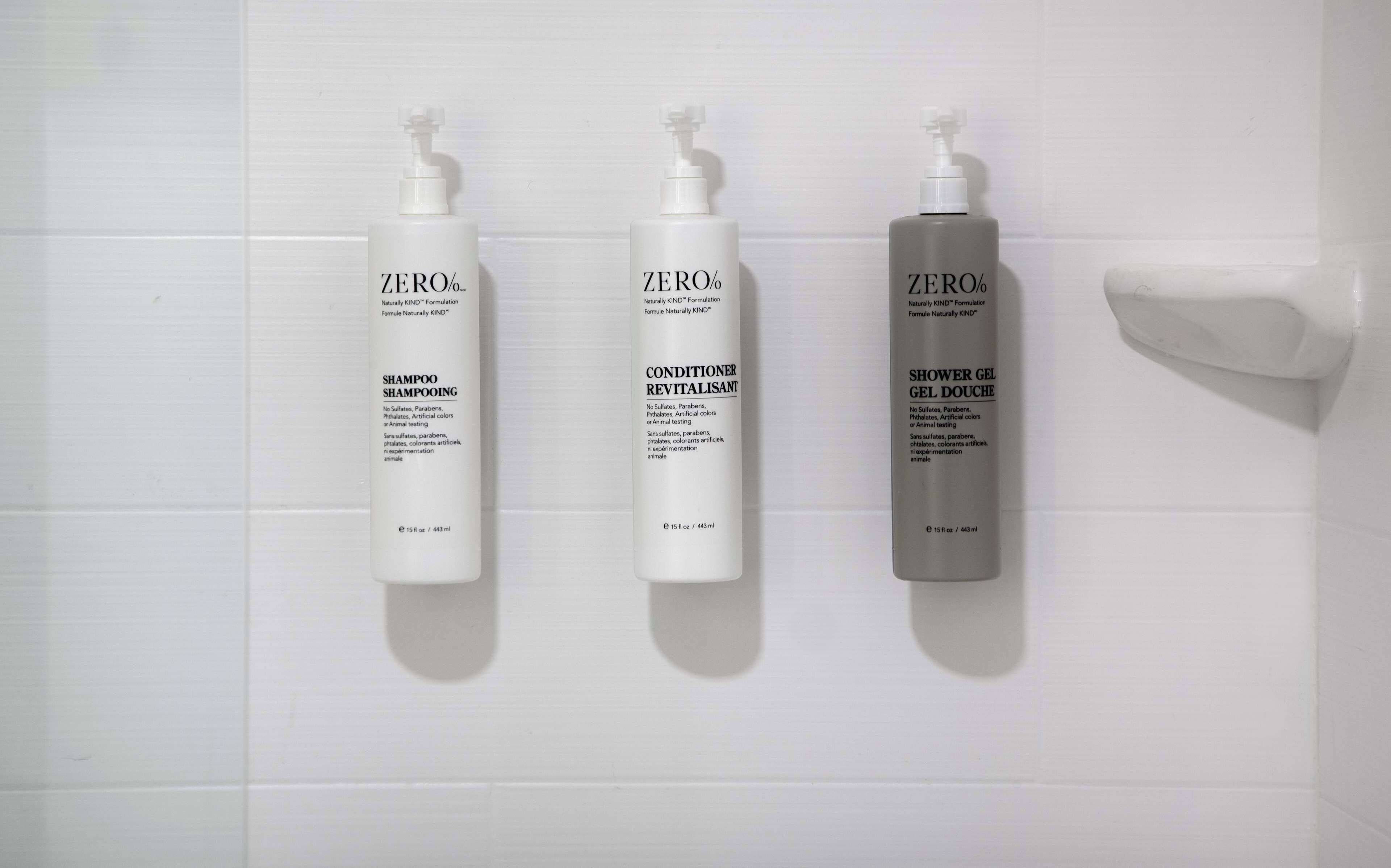 Three bottles of Zero% shampoo, conditioner, and shower gel on a white tiled wall.