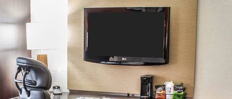 Una camera d'albergo con televisione a schermo piatto fissata al muro, una scrivania con una sedia, una macchina del caffè e un piccolo cesto con degli snack.